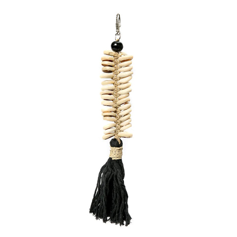 A handmade keychain featuring a vertical arrangement of small beige seashells intricately woven together, topped with a black bead. A thick black cotton tassel hangs at the bottom, and a metal clasp is attached at the top.