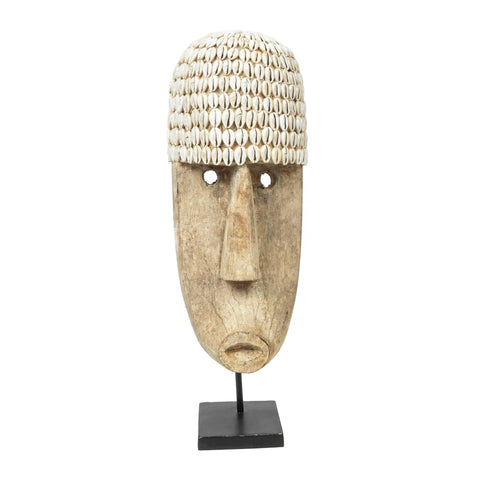 A wooden tribal mask with a long face and hollow eyes, adorned with hand-placed shells forming a headdress, embodies ethnic beauty. Perfect for enriching any home interior, it is elegantly displayed on a black stand.