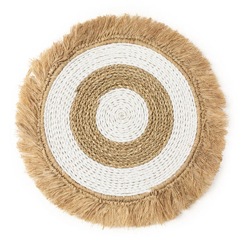 A round, woven seagrass placemat with concentric circles in natural beige and white, featuring a fringed edge, perfect for adding a touch of boho interior style.