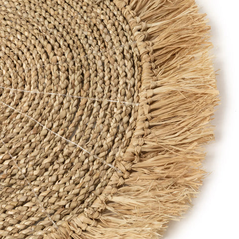 Close-up of a round, woven jute fiber mat with a detailed braided pattern, reminiscent of raffia placemats. The fringe border made of natural fibers adds texture and rustic charm, perfect for enhancing any boho interior.