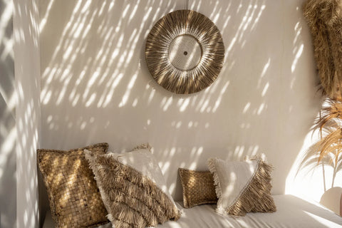 A cozy interior with a beige couch adorned with bohemian cushion covers against a wall. A circular, straw wall decoration casts soft shadows. Sunlight filters through, creating an inviting, coastal vibe.