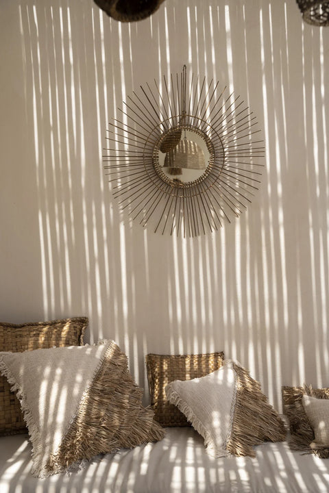 Sunlight casts striped shadows on a wall with a decorative round mirror featuring a spiked frame. Below, several textured cushion covers in earthy tones are arranged on a white surface, creating a warm and inviting bohemian atmosphere.