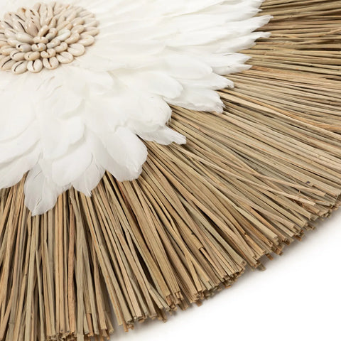 Close-up of an Alang Alang Feather Juju with white feather-like petals encircling a central cluster of light-colored seeds, bordered by cowrie shells and natural dried grass sticks radiating outward.