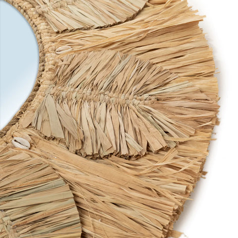 Close-up of a decorative mirror framed with woven natural raffia fibers and tassels. The texture is earthy and rustic, offering an organic look. Small cowrie shells are seamlessly integrated into the design for added charm.