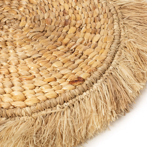 Close-up of a round woven raffia placemat, featuring a braided pattern and fringed edges. The texture is highlighted, showcasing various shades of beige and brown, perfect for adding a touch of boho interior charm.