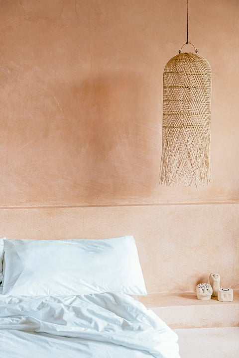 A minimalist bedroom showcases a bed with white bedding against a peach-colored wall. A woven rattan lamp pendant light elegantly hangs from the ceiling, adding texture and warmth. Two small decorative items are placed on a shelf beside the bed.