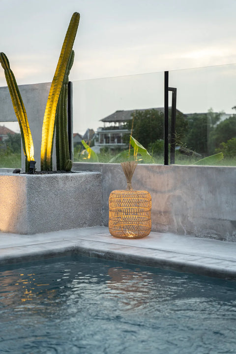 Modern outdoor pool area with a woven rattan lamp glowing beside a tall potted cactus. Glass railing offers a view of greenery and a distant house. The sky above is overcast, creating a calm atmosphere.