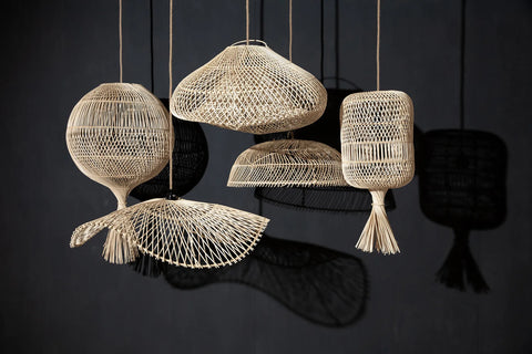 A group of intricately woven boho rattan pendant lamps hangs against a dark background. The multi-purpose lamps vary in shape and size, casting interesting shadows on the wall.