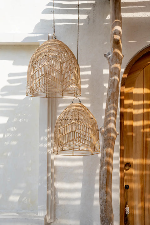 Two handcrafted rattan pendant lights hang from the ceiling, casting shadows on a white textured wall. A curved wooden door is partially visible on the right, with a tall, slender tree trunk beside it, adding a natural element to the minimalist setting.