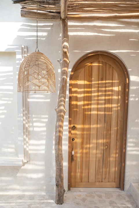 A rustic wooden door with a curved top is set in a white stucco wall. A natural wood beam supports a thatched roof, casting shadows. A handcrafted rattan pendant light hangs nearby, enhancing the earthy, natural ambiance.