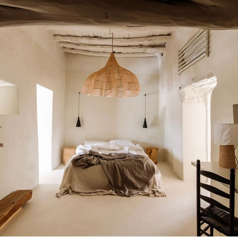 A rustic bedroom featuring a cozy bed with beige linens is embraced by white stucco walls and wooden beams on the ceiling. An XXL rattan pendant lamp hangs above, exuding boho chic vibes, while a simple chair sits near a small wooden table, all softly illuminated by natural lighting.