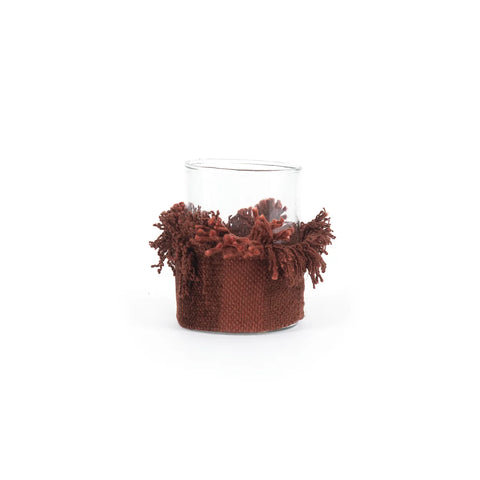 A charming glass candle holder with a textured, hand-knitted brown woven sleeve featuring tassels adorning the upper part, wrapped elegantly around the center.