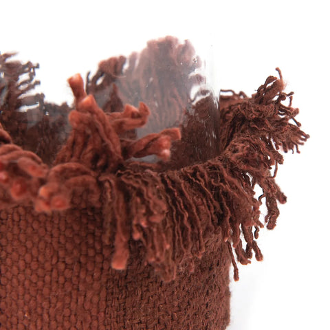 Close-up of a glass bottle wrapped in a textured, hand-knitted brown cozy with fringe at the top. The yarn appears soft and inviting against the plain white background.