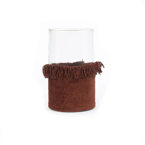 A clear glass vase with a brown, textured fabric cover around the base and fringe detailing at the top. Reminiscent of velvet jackets, the fringe adds a decorative touch to the minimalist design. The background is plain white.