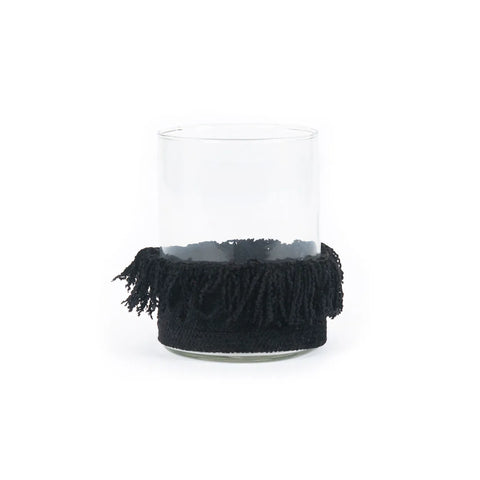 A clear glass adorned with a black, hand knitted fringed fabric ring around its bottom half sits against a plain white background.