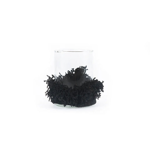 A clear glass candle holder with a black textured ornamental wrap around the base, resembling intricate lace or fringe, evokes the cozy charm of knitted cotton. Its elegant design stands out against a plain white background.