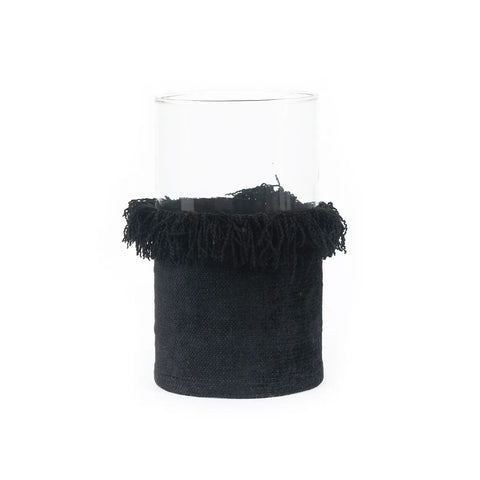 A transparent cylindrical glass vase partially wrapped in a hand-knitted black fabric with a fringe around the top, resembling elegant candle holders.