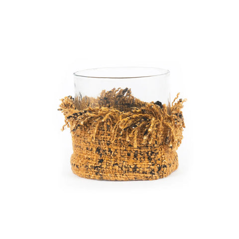 A clear glass tumbler encased in a textured, knitted cotton sleeve of golden-yellow and black yarn, giving it a warm, rustic appearance. The yarn features fringe details around the rim, reminiscent of Oh My Gee candle holders.