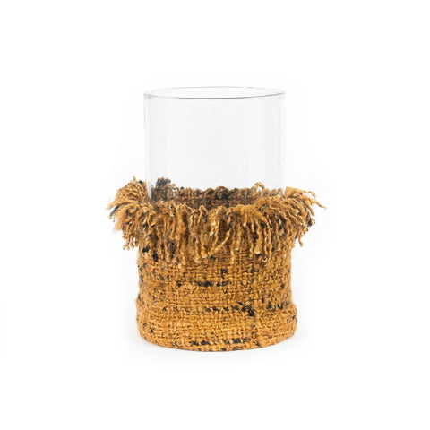 A clear glass vase with a decorative, textured brown knitted cover wrapped around its lower half. This home decor piece has a fringed top edge, giving it a cozy and rustic appearance, reminiscent of charming glass jackets.