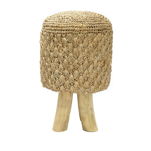 A woven rattan and raffia stool with a textured, dome-shaped seat and two teak wood legs, resembling a mushroom. The natural fibers create an intricate pattern, evoking a boho summer vibe. Set against a plain white background.