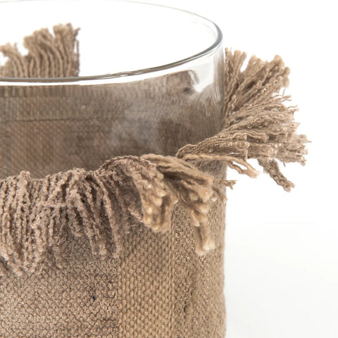 Close-up of a glass vase wrapped in a textured, burlap-like fabric with frayed edges, evoking a rustic, hand-knitted charm. The fibers extend outward unevenly, providing a distinctive, handcrafted appearance reminiscent of cozy candle holders.