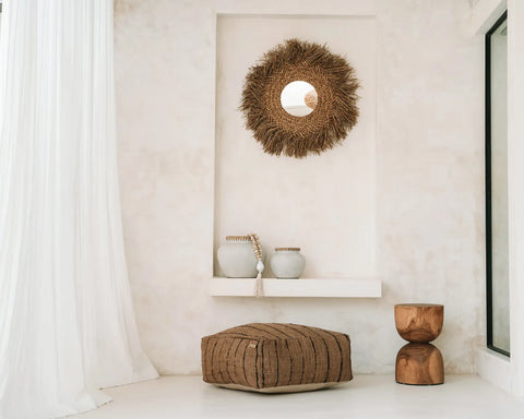 Minimalist interior featuring a textured cotton pouffe, a wooden stool, and a shelf displaying two vases. A round straw mirror hangs above on a light beige wall. Soft, flowing white curtains partially cover a window on the left, completing this serene home accessory setup.