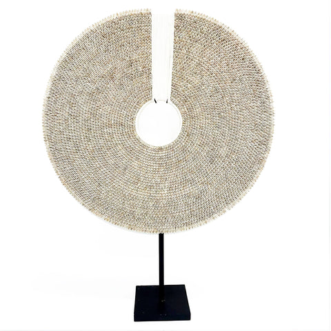 A large, round sculpture with a hollow center, resembling a disk, evokes a coastal feel. Mounted on a thin black stand, it features intricately overlapping shells and a slight opening on top. This statement piece's textured surface captivates with small, tightly packed elements.