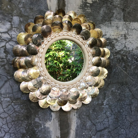 A coastal wall mirror with a decorative frame of overlapping, metallic scallop-shaped discs is mounted on a textured gray concrete wall. The reflection of green bamboo leaves adds a touch of boho chic styling to the mirror's surface.