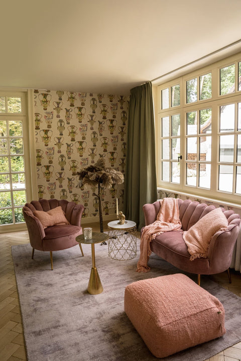 A cozy living room features a pink armchair and sofa, both adorned with soft cushions and a throw. A small white round side table with a brass base sits between them, alongside a fluffy decorative piece. Large windows offer a view of greenery outside.