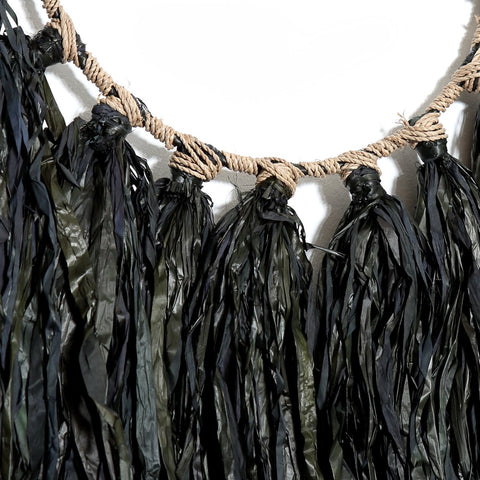 A close-up of a bohemian wall hanger showcasing dark green and black tassels crafted from raffia, resembling dried leaves or seaweed, attached to a braided rope. Set against a plain white wall, this piece adds an earthy touch.