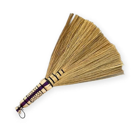 This Boho-inspired hand broom, reminiscent of Indonesian hand sweepers, is crafted from natural straw and features a woven handle with black and purple accents. Its straw bristles fan out beautifully, and there's a loop at the end for easy hanging.