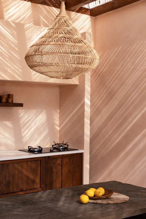 A stylish kitchen with warm tones features a large woven rattan lamp shade casting intricate shadows. A rustic wooden countertop holds a wooden bowl with lemons. The background reveals a gas stovetop and wooden shelves, as sunlight filters through slatted blinds.