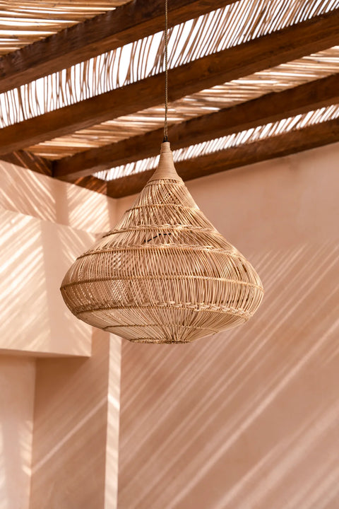 A woven rattan lamp shade hangs from a wooden pergola. Sunlight filters through the slats, casting intricate shadows on the peach-colored wall and floor beneath. The ambiance is warm and rustic.