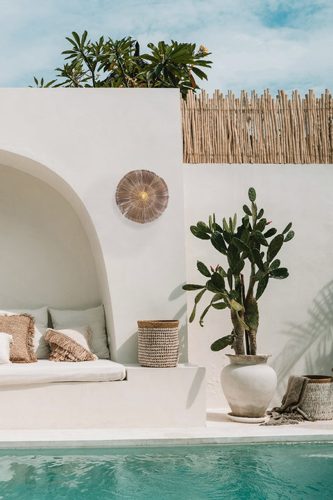 A minimalist outdoor space features a white stucco wall with a built-in bench adorned with cushions in neutral tones. A Chi-Chi wall lamp adds elegance, while a large cactus by the swimming pool and a wicker basket enhance the serene setting.