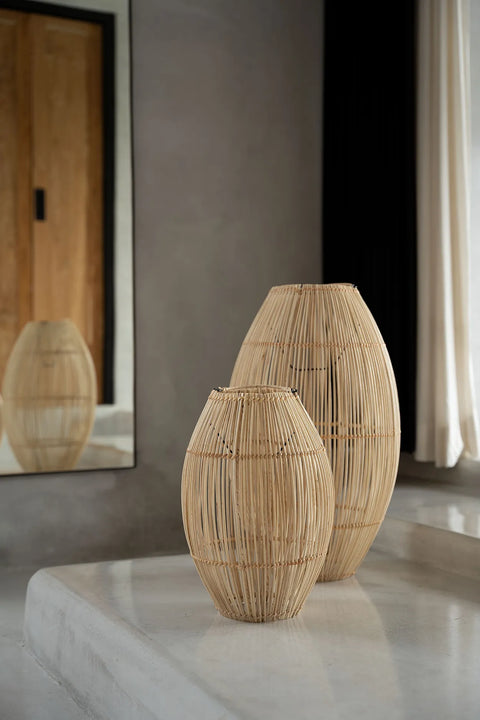 Two woven wicker vases of different sizes sit on a concrete ledge in a minimalist room. A large mirror and cozy stripes of light from a rattan lamp create an inviting atmosphere, casting soft reflections on the vases and curtain in the background.