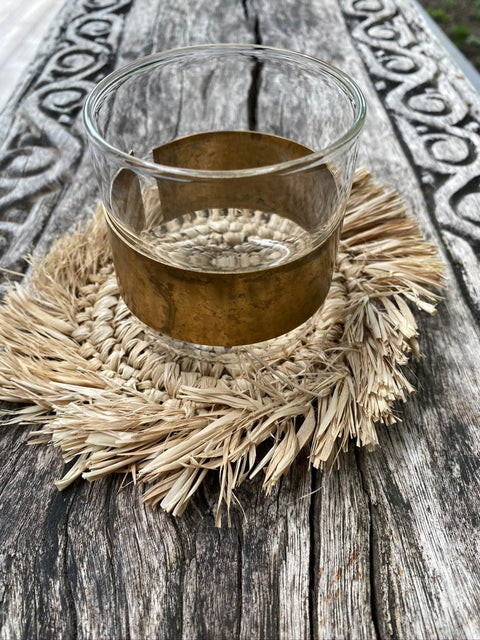 A glass with a gold band rests on a natural wicker coaster atop a weathered wooden surface. The intricate carvings on the wood provide a rustic background.