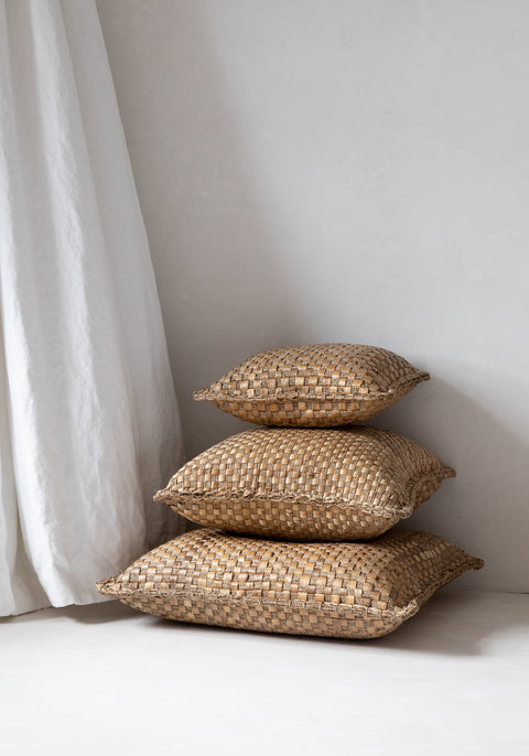 In the corner on a light floor, three stacked woven pillows exude a boho vibe as soft furnishings. Their natural, textured look contrasts beautifully against the smooth white wall and a flowing curtain.