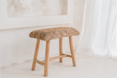 A raffia stool with a woven seat is placed in a minimalist room with light-colored walls and a sheer white curtain, exuding a subtle summer vibe.